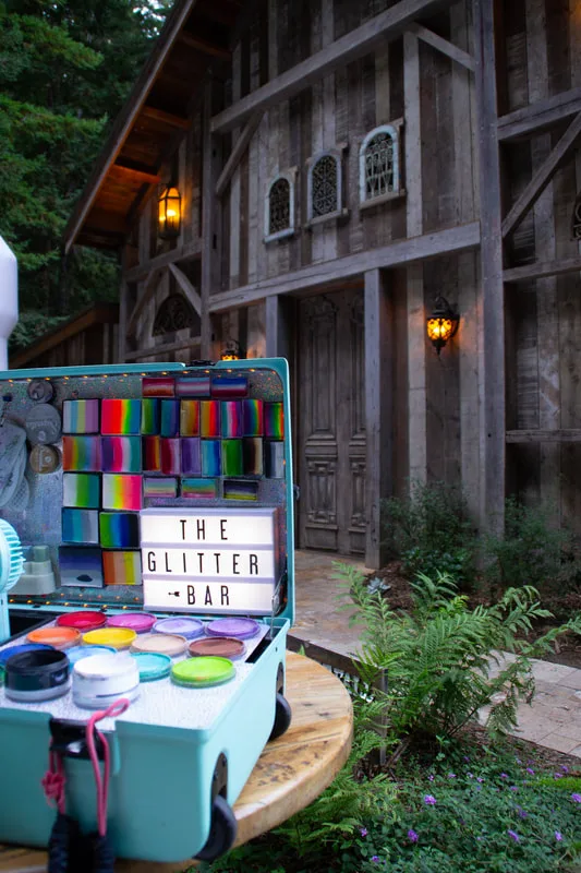 A photo of the glitter bar set up for a wedding welcome party in Ben Lomand, California. The glitter bar features a tiffany blue suitcase filled with rainbow face painting cakes and glitter and a light up sign advertising the Glitter Bar