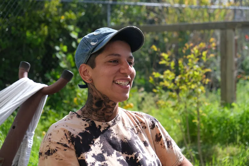 A smiling person with a medium skin tone models a realistic airbrush tattoo of a skull and bones on their neck