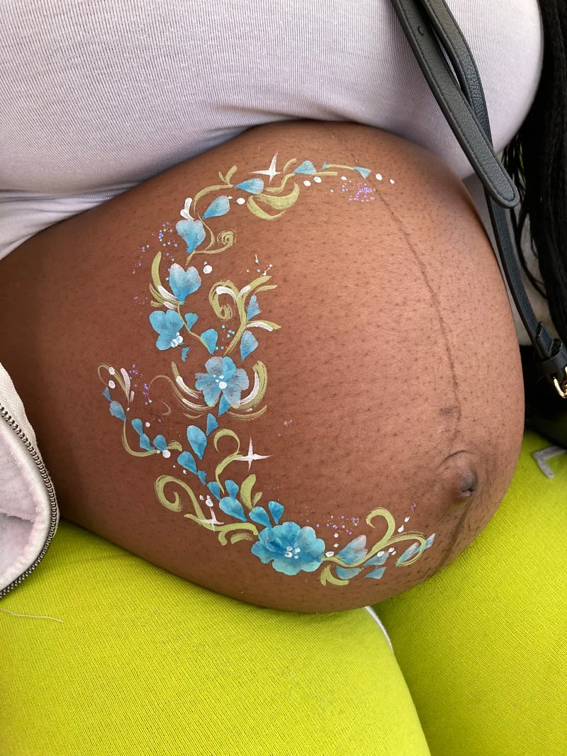 A pregnant woman in a white shirt and yellow pants has blue flowers painted on her belly