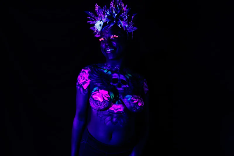 A young, smiling black woman in San Francisco, Ca, model's a UV reactive body painting and blacklight reactive eyelashes. The design splashes across her chest, a skull in the center with wing's spreading out following her collar bone. Glowing pink roses and white mandala's surround the design. She also sports a spikey gold and black floral crown.