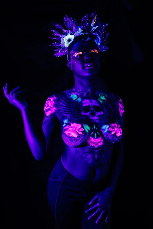 A young black woman in San Francisco, Ca, model's a UV reactive body painting and blacklight reactive eyelashes. The design splashes across her chest, a skull in the center with wing's spreading out following her collar bone. Glowing pink roses and white mandala's surround the design. She also sports a spikey gold and black floral crown.