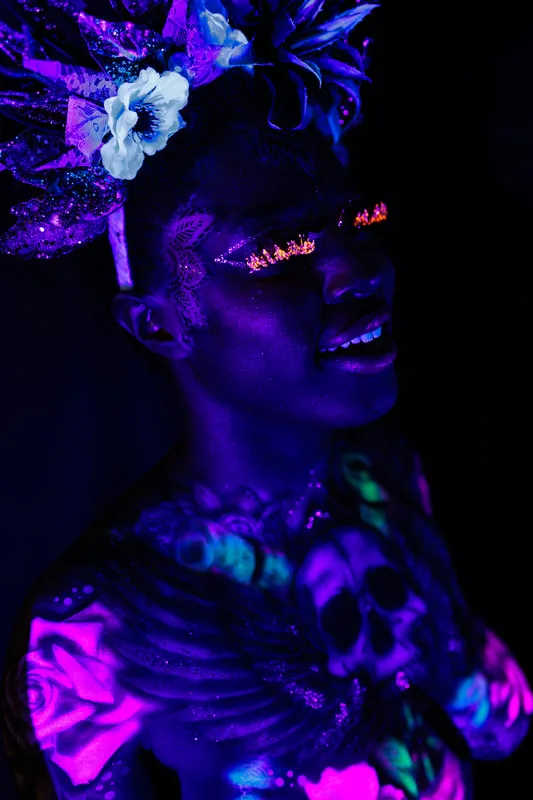 A young, smiling black woman in San Francisco, Ca, model's a UV reactive body painting and blacklight reactive eyelashes. The design splashes across her chest, a skull in the center with wing's spreading out following her collar bone. Glowing pink roses and white mandala's surround the design. She also sports a spikey gold and black floral crown.