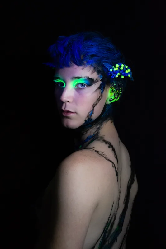 A young white woman in Oakland, Ca is wearing UV reactive body paint and blacklight activated makeup. She resembles a fairy with a head piece and pointed ear cuff's, but the design in inspired by fungus and rot. Black tendrils sweep up around her neck like a necklace and top. Green berries and blue plant matter glow in the dark.
