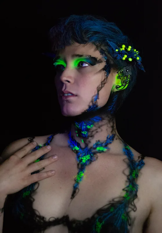 A young white woman in Oakland, Ca is wearing UV reactive body paint and blacklight activated makeup. She resembles a fairy with a head piece and pointed ear cuff's, but the design in inspired by fungus and rot. Black tendrils sweep up around her neck like a necklace and top. Green berries and blue plant matter glow in the dark.