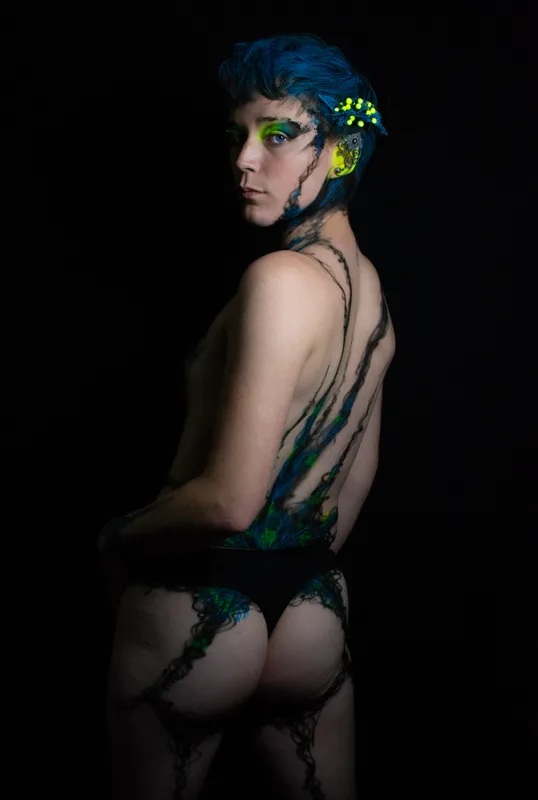 A young white woman in Oakland, Ca is wearing UV reactive body paint and blacklight activated makeup. She resembles a fairy with a head piece and pointed ear cuff's, but the design in inspired by fungus and rot. Black tendrils sweep up around her neck like a necklace and top. Green berries and blue plant matter glow in the dark.