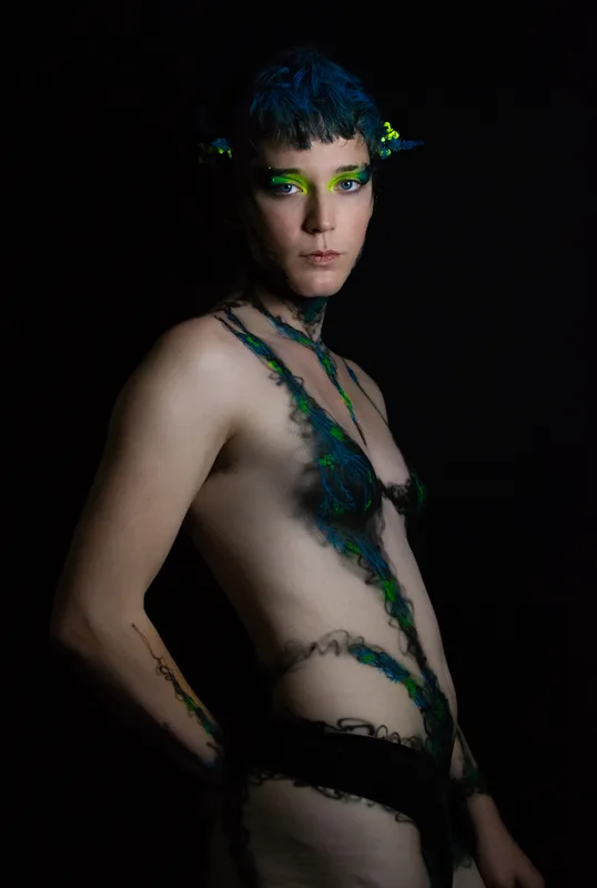 A young white woman in Oakland, Ca is wearing UV reactive body paint and blacklight activated makeup. She resembles a fairy with a head piece and pointed ear cuff's, but the design in inspired by fungus and rot. Black tendrils sweep up around her neck like a necklace and top. Green berries and blue plant matter glow in the dark.