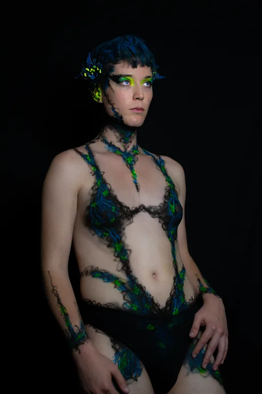A young white woman in Oakland, Ca is wearing UV reactive body paint and blacklight activated makeup. She resembles a fairy with a head piece and pointed ear cuff's, but the design in inspired by fungus and rot. Black tendrils sweep up around her neck like a necklace and top. Green berries and blue plant matter glow in the dark.
