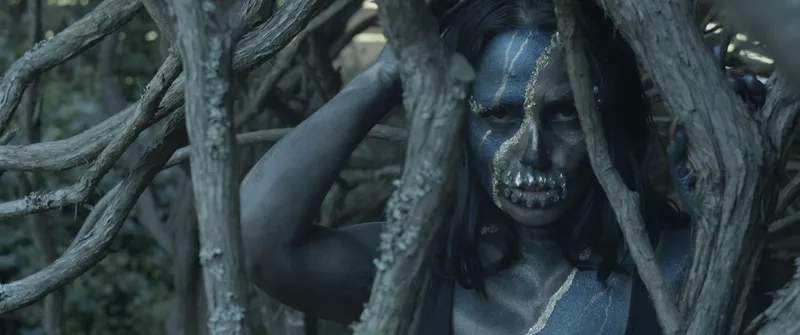 A young woman wears blue and gold skull face and body painting with diamond teeth while standing in a tree grove, San Francisco