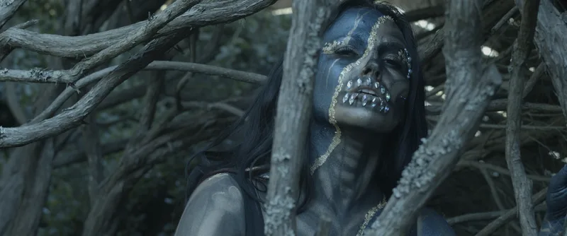 A young woman wears blue and gold skull Face and body painting with diamond teeth while standing in a tree grove, San Francisco